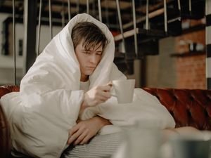 Man wrapped in a blanket sipping hot drink while seated on a cozy sofa indoors.